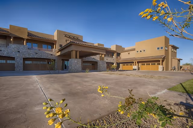 Exterior view of a modern senior living facility building with stone and stucco walls, multiple garage doors, and a covered entrance under a clear blue sky. Yellow flowering plants are visible in the foreground.
