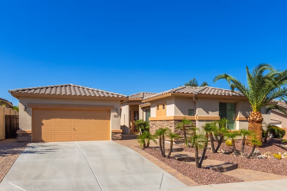 Single-story tan house with a two-car garage, desert landscaping, and blue sky.