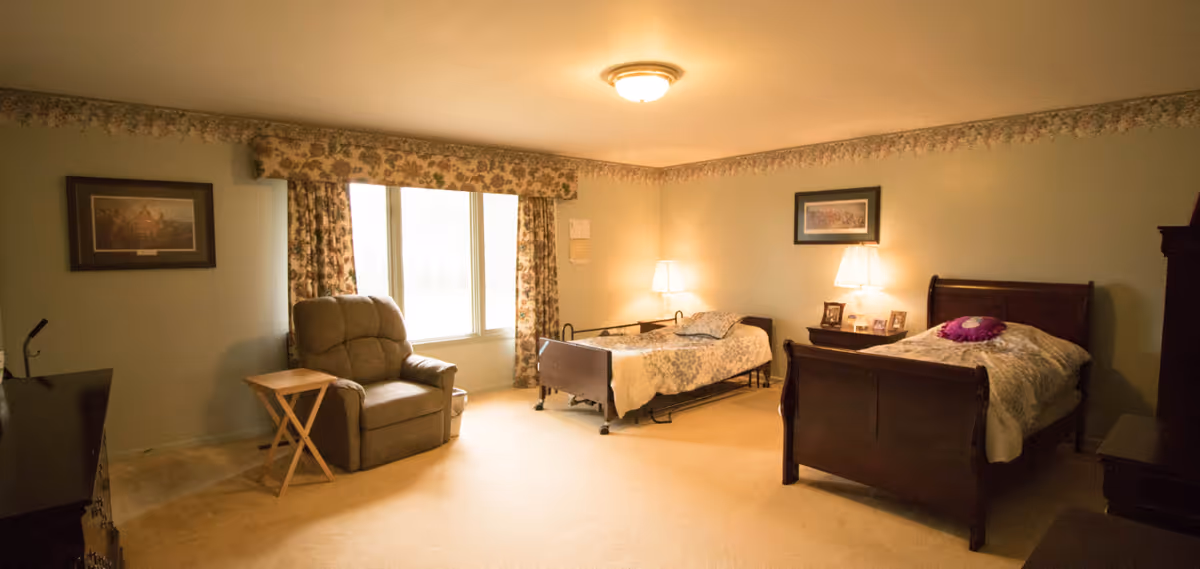 Two beds, a recliner, lamps, and nightstands sit in a cozy carpeted senior living bedroom.