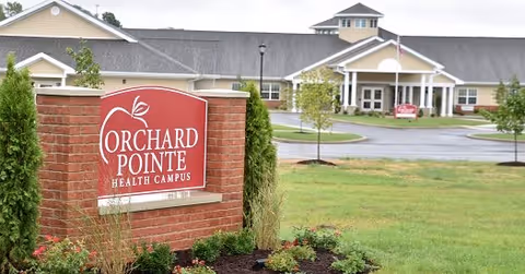Brick sign reading "Orchard Pointe Health Campus" in front of the facility building and landscaped lawn.