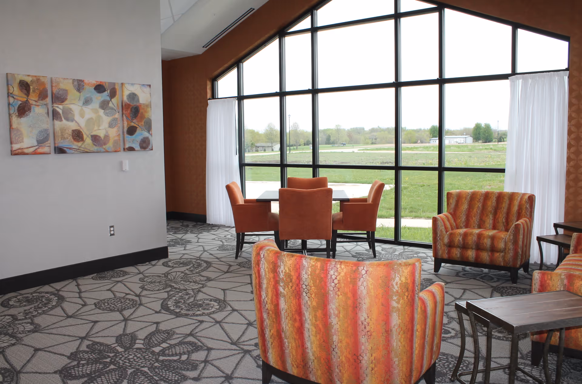 A bright room with large floor-to-ceiling windows overlooking a green outdoor area. The room features patterned carpet, orange and red striped armchairs, a small wooden table, and a dining table with four orange chairs. There is a three-panel abstract painting on the wall and white curtains partially drawn to the sides of the windows.