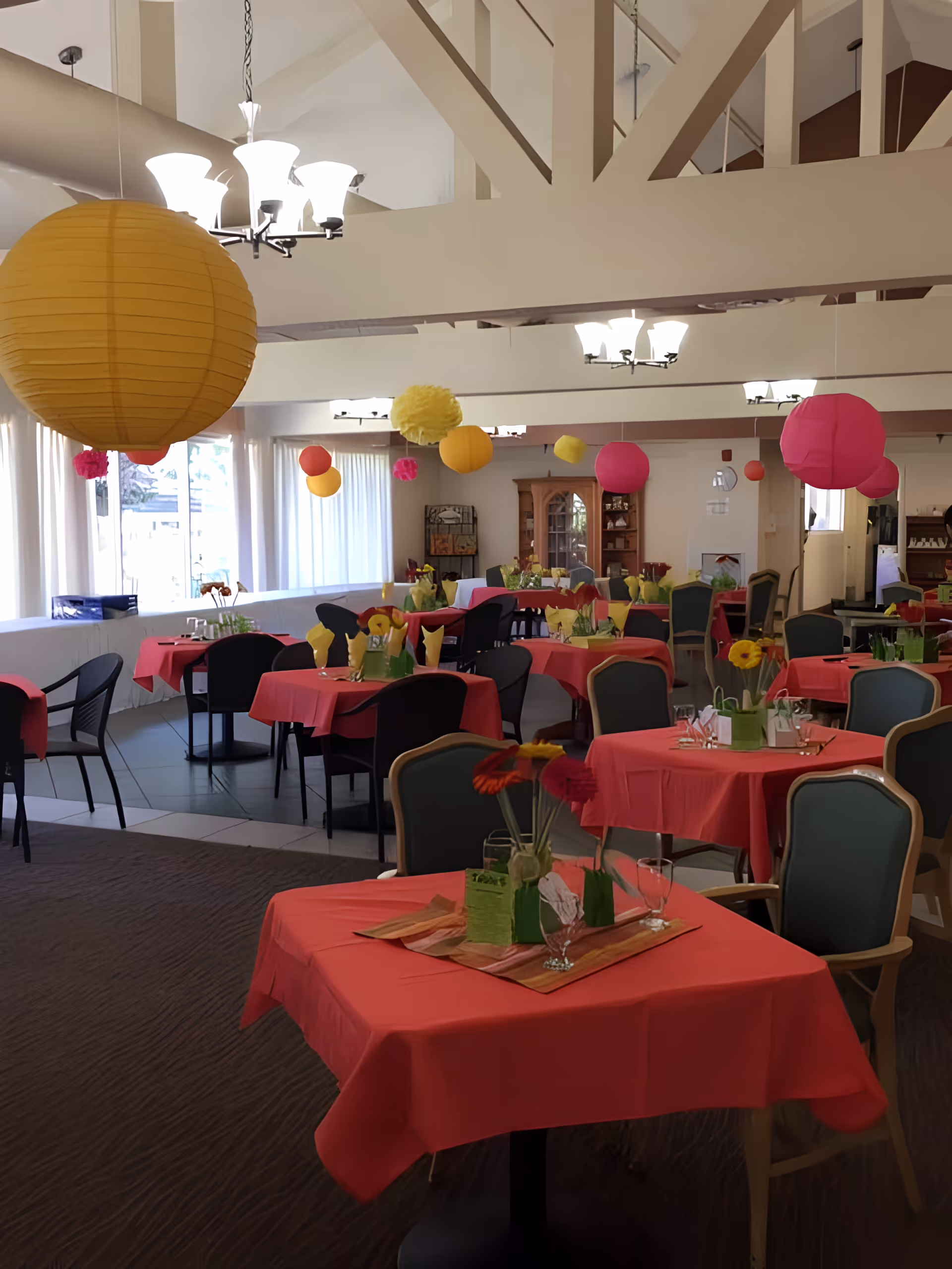 A dining room decorated with red tablecloths and colorful paper lanterns hanging from the ceiling. Tables are set with glasses, napkins, and flower centerpieces. The room has large windows with white curtains and a high ceiling with exposed beams.