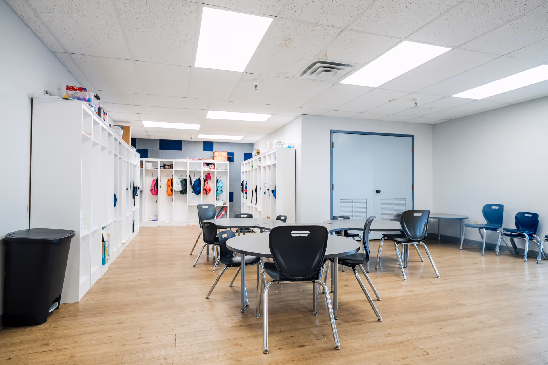 A bright indoor activity room with lockers, tables, chairs, and blue doors.