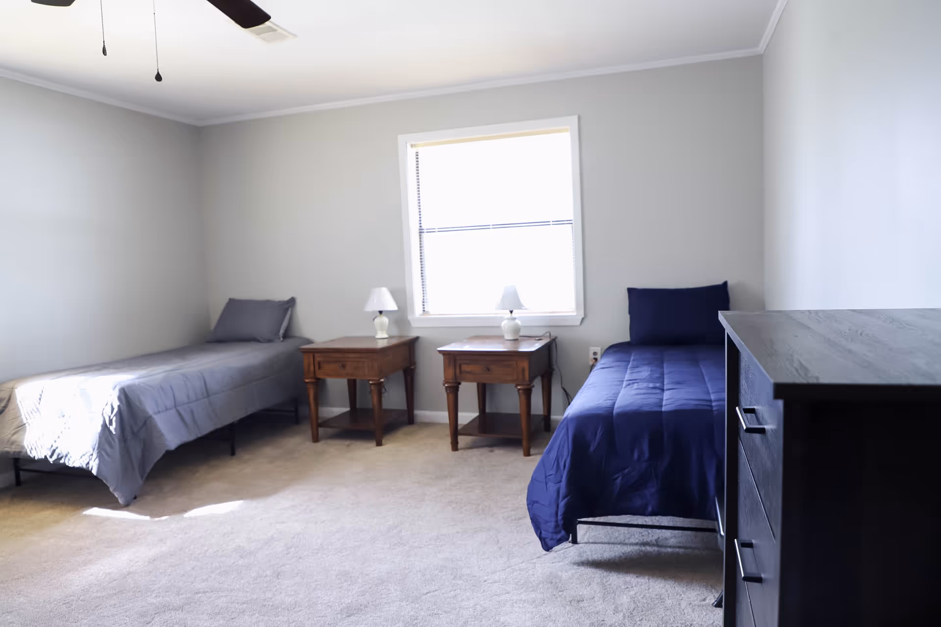 A simple bedroom with two single beds, one with gray bedding and the other with dark blue bedding. Between the beds are two wooden nightstands, each with a small white lamp. There is a window centered on the wall behind the nightstands, and a dark wooden dresser is visible on the right side of the image. The walls are painted light gray and the floor is carpeted.
