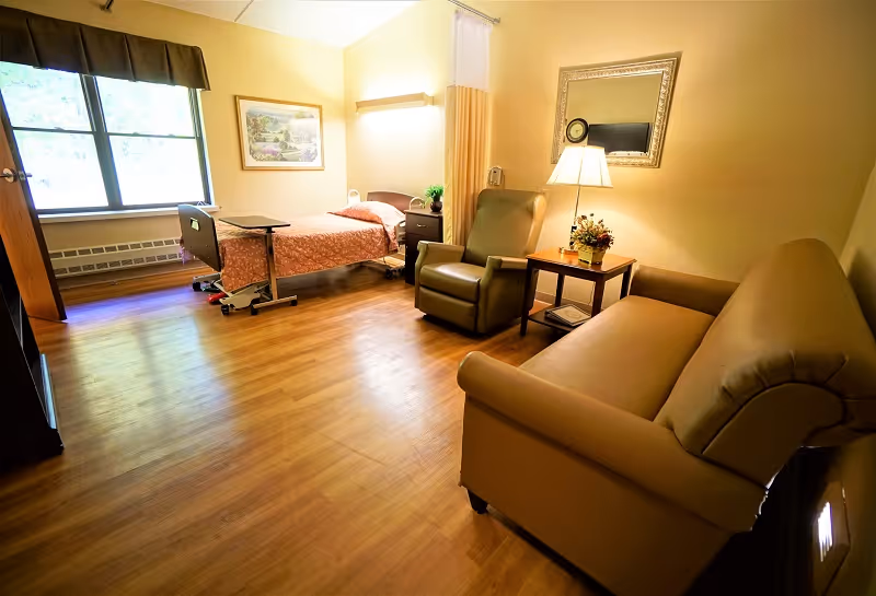Furnished resident bedroom with an adjustable single bed, recliner, sofa, side table with lamp, and a large window.