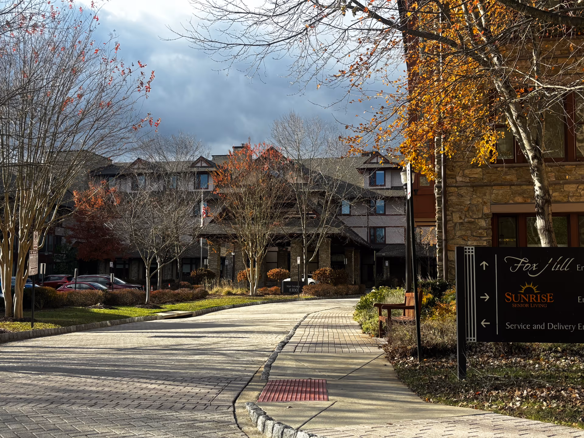 Exterior view of Sunrise at Fox Hill senior living facility showing a paved driveway leading to the main entrance of a multi-story building with stone and wood accents. Trees with autumn leaves and some bare branches surround the area under a cloudy sky. A sign near the sidewalk displays directions and the Sunrise Senior Living logo.