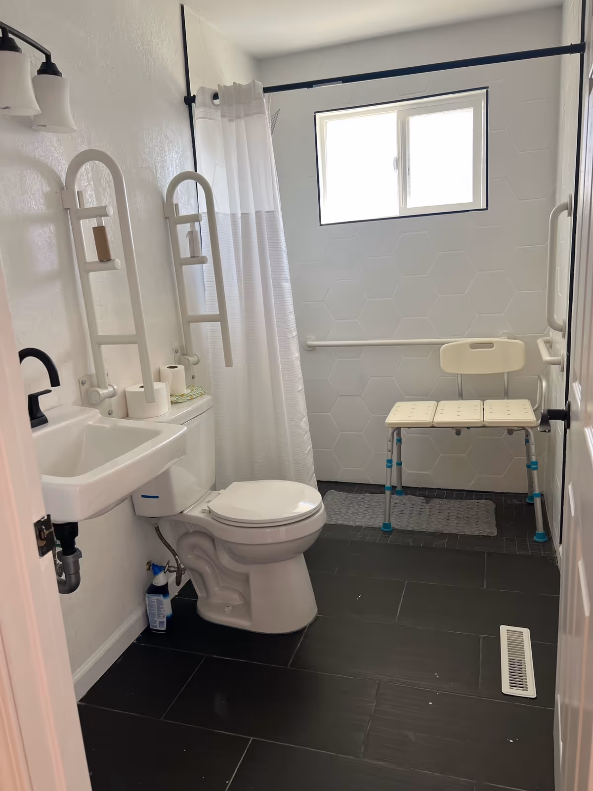 Accessible bathroom with sink, toilet, grab bars, and a shower chair in a senior facility.