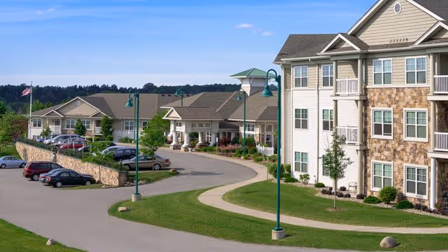 Exterior view of a senior living facility with multiple buildings, a curved driveway, parked cars, green lawns, and street lamps under a clear blue sky.