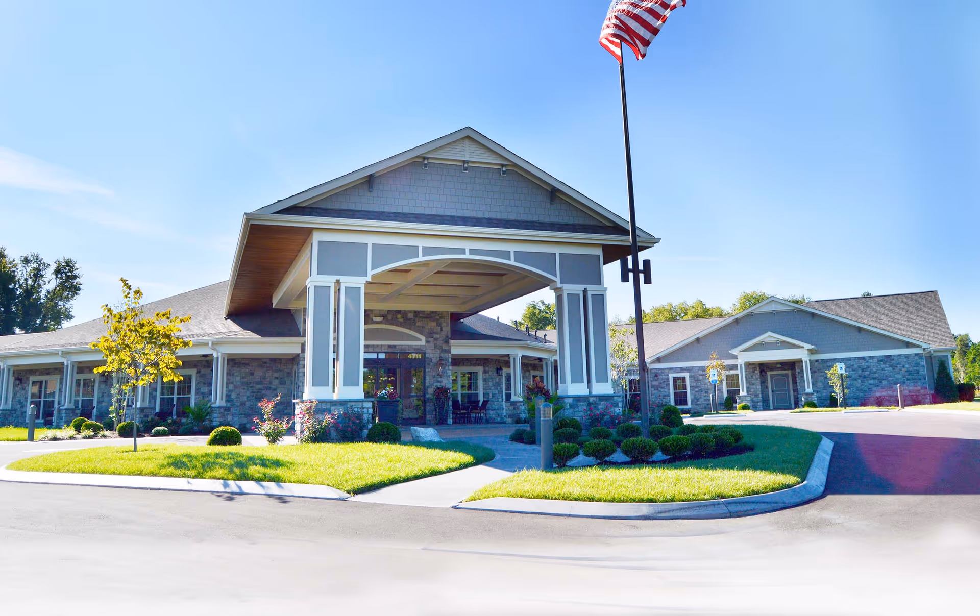 Front entrance of a single-story senior living facility with a large covered porte-cochere, landscaped lawns, and an American flag.