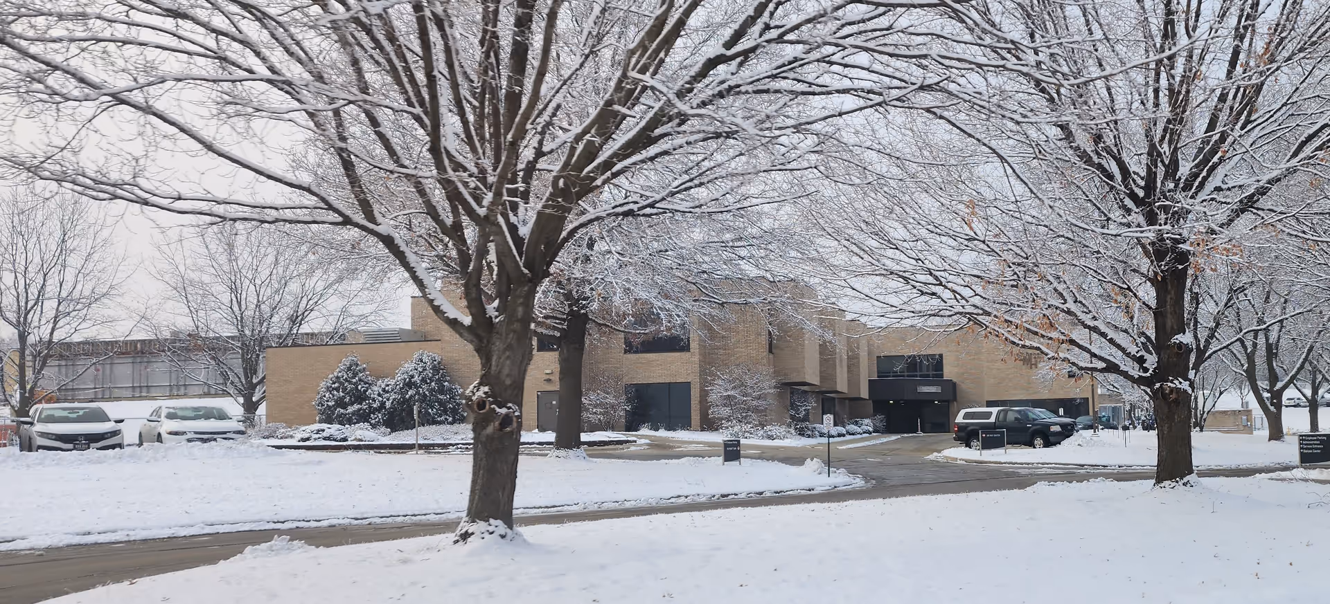 Snow covers a modern medical center exterior with trees and parked cars along the driveway.