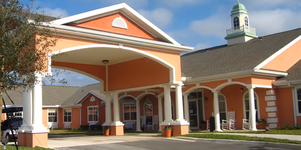 Exterior view of a senior living facility building with peach-colored walls, white columns, and a covered entrance with rocking chairs on the porch under a partly cloudy sky.