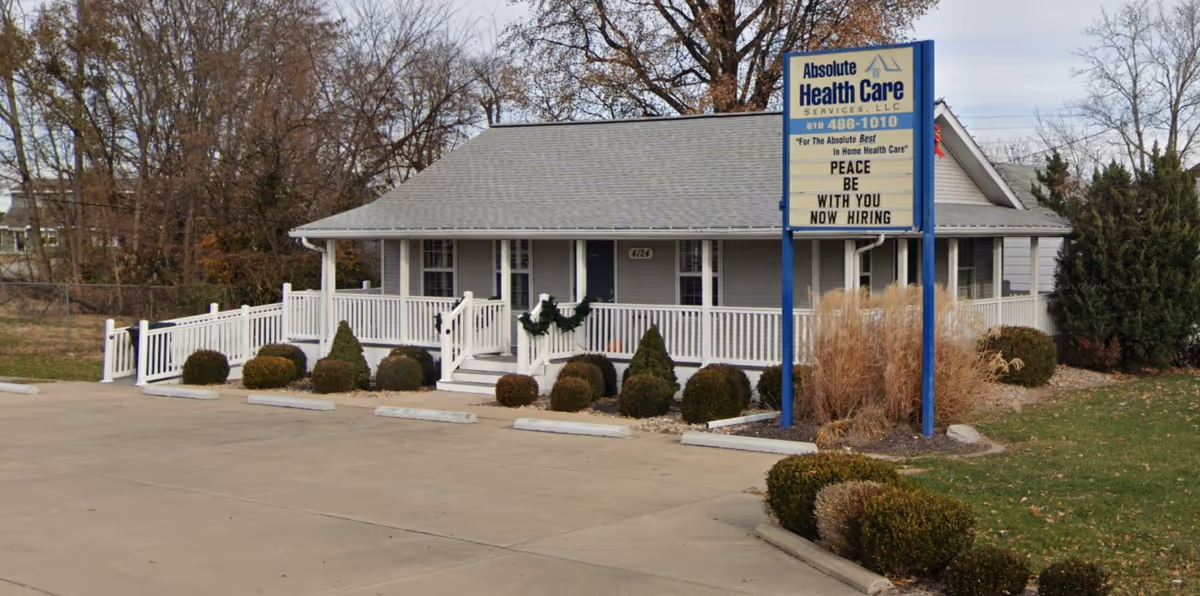 Single-story care facility with a blue-and-white front porch, sign, and trimmed shrubs.