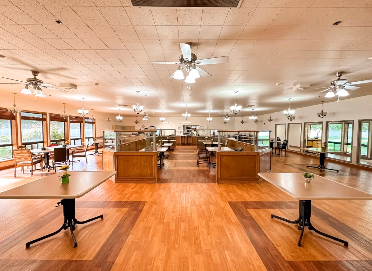 Spacious dining hall with multiple tables, partitions, and ceiling fans in a senior facility.