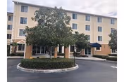 Three-story beige senior living building with entry, curved driveway, and landscaped circular island.