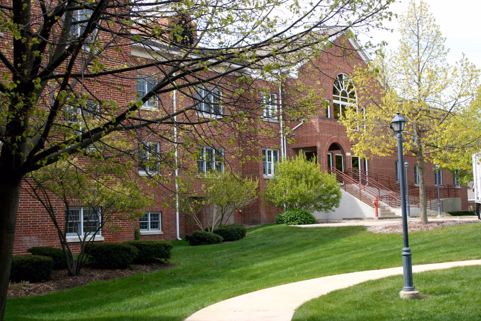 Exterior view of a red brick building with multiple windows, surrounded by green grass, trees with budding leaves, and a curved concrete pathway leading to the entrance. A lamppost is visible near the pathway.