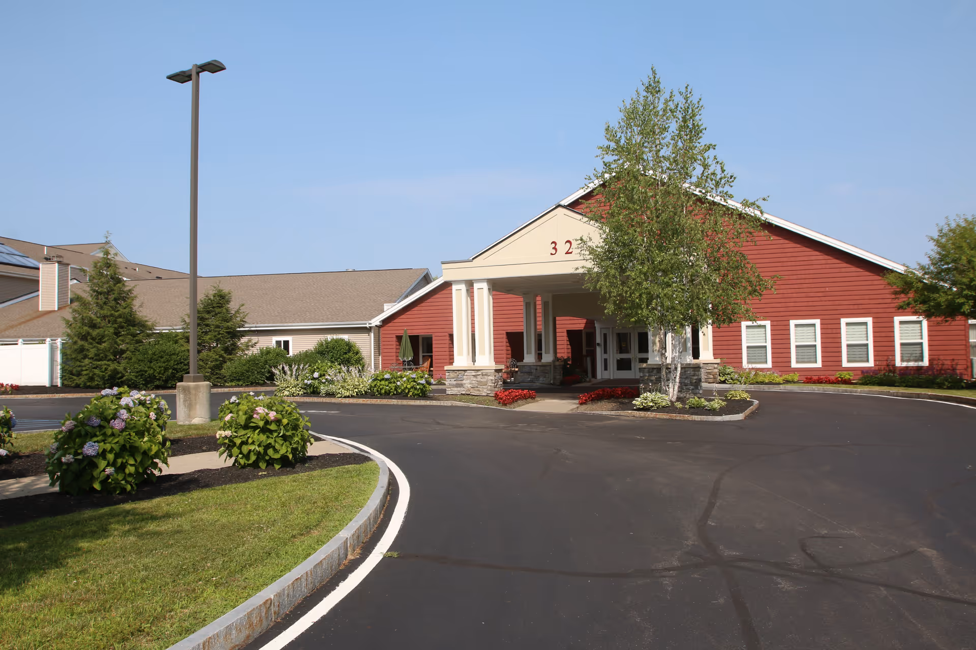 Front entrance of a red senior living building with a covered portico, circular driveway, and landscaped beds.