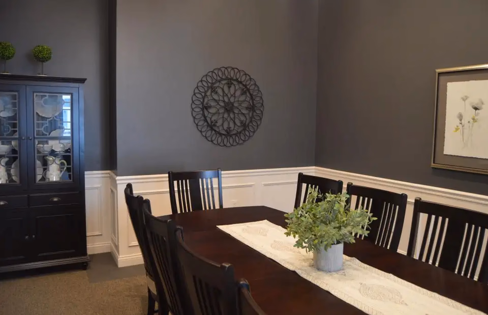 Dark dining room with a long wooden table, chairs, a plant centerpiece, and wall decor.
