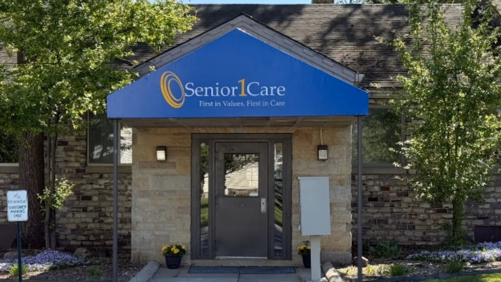 Front entrance of Senior1Care facility with blue canopy, stone walls, logo, and surrounding trees.