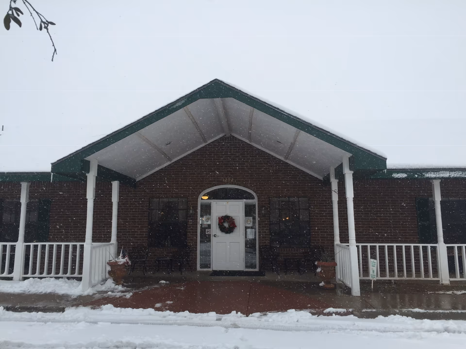 Front entrance of a brick building with a covered porch, a white door decorated with a wreath, and snow on the ground.