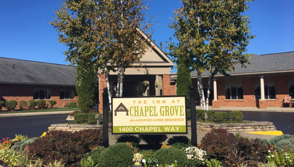 Entrance sign for The Inn at Chapel Grove (1400 Chapel Way) with landscaped gardens and a single-story brick assisted living building behind it under a blue sky.