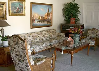 A living room with floral upholstered sofa and chairs, a wooden coffee table with decorative items, a potted plant, framed paintings on the wall, and a small organ in the corner.