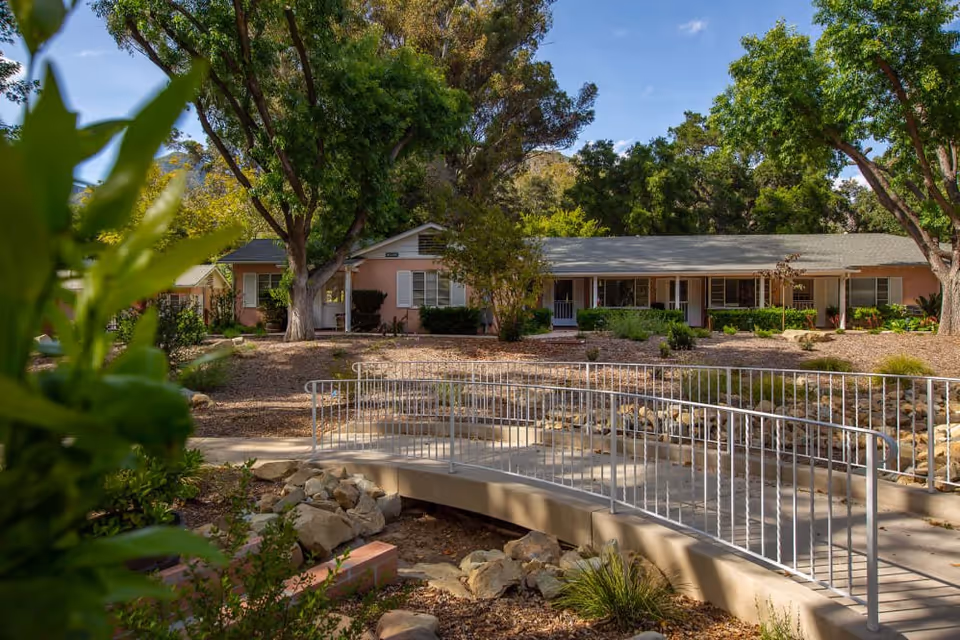 A single-story building with a gray roof and light pink exterior surrounded by trees and greenery. In the foreground, there is a curved pedestrian bridge with metal railings crossing over a dry rocky creek bed. The sky is clear and blue.
