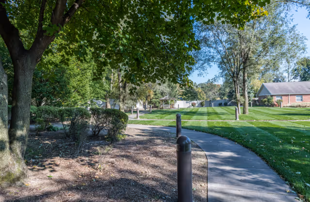 A paved winding sidewalk along green lawns and trees by a residential building.