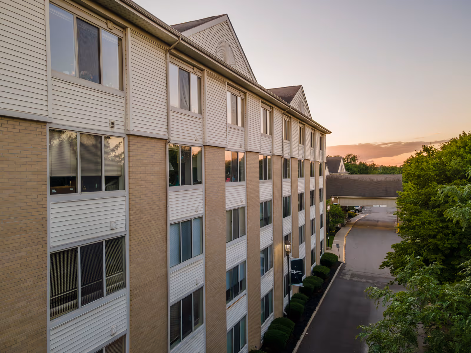 Exterior view of a multi-story senior living building with rows of windows, a driveway and trees at sunset.