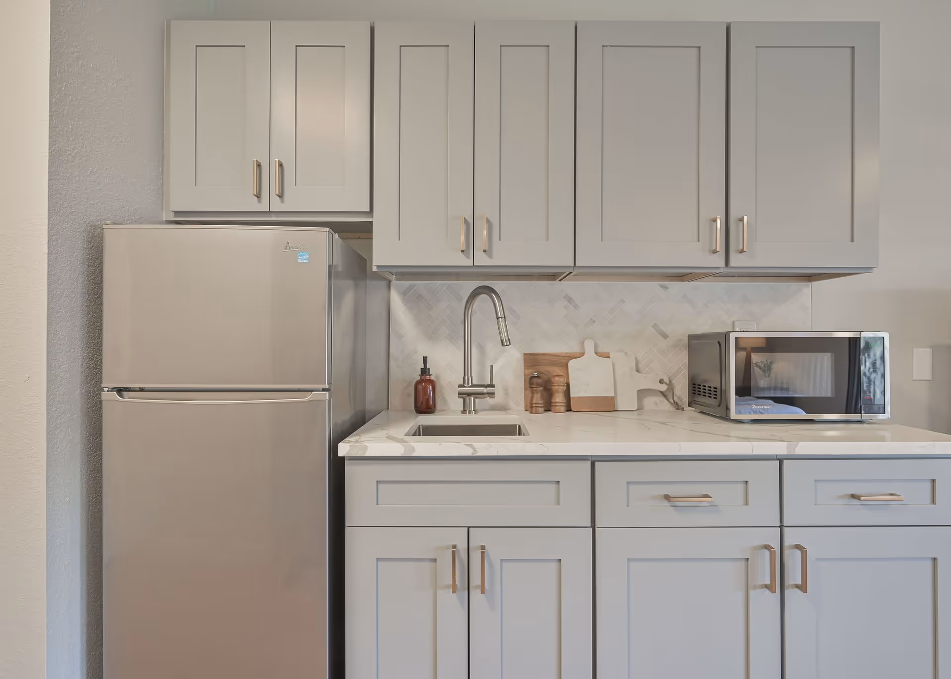 Modern kitchen area with light gray cabinets, a stainless steel refrigerator, a sink with a curved faucet, a soap dispenser, two cutting boards, salt and pepper shakers, and a microwave on a white marble countertop.