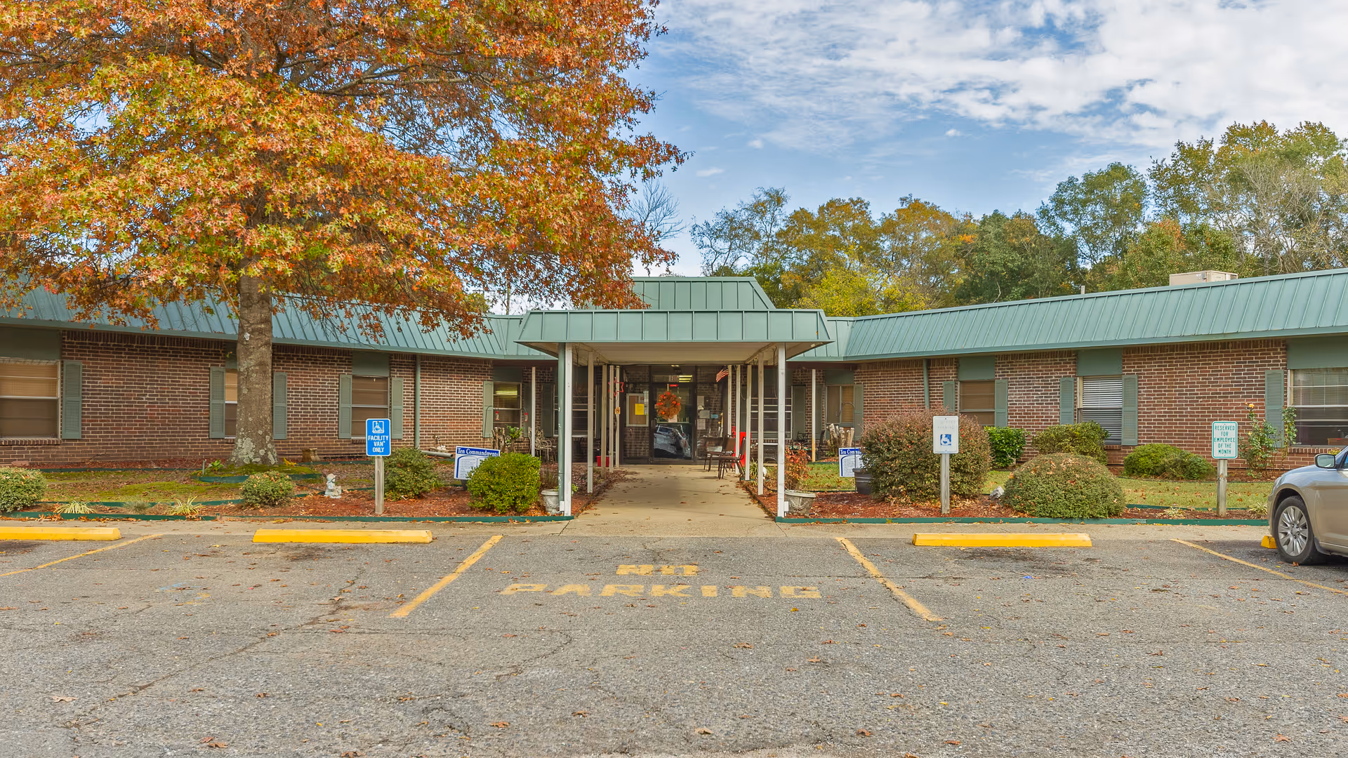Front exterior view of a single-story brick building with a green metal roof, a covered entrance walkway, and autumn-colored trees. There are parking spaces and handicap parking signs in front of the building.