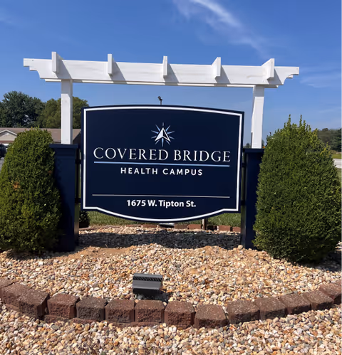 Outdoor entrance sign that reads "Covered Bridge Health Campus, 1675 W. Tipton St." set in a rock bed with shrubs and a white pergola against a blue sky.