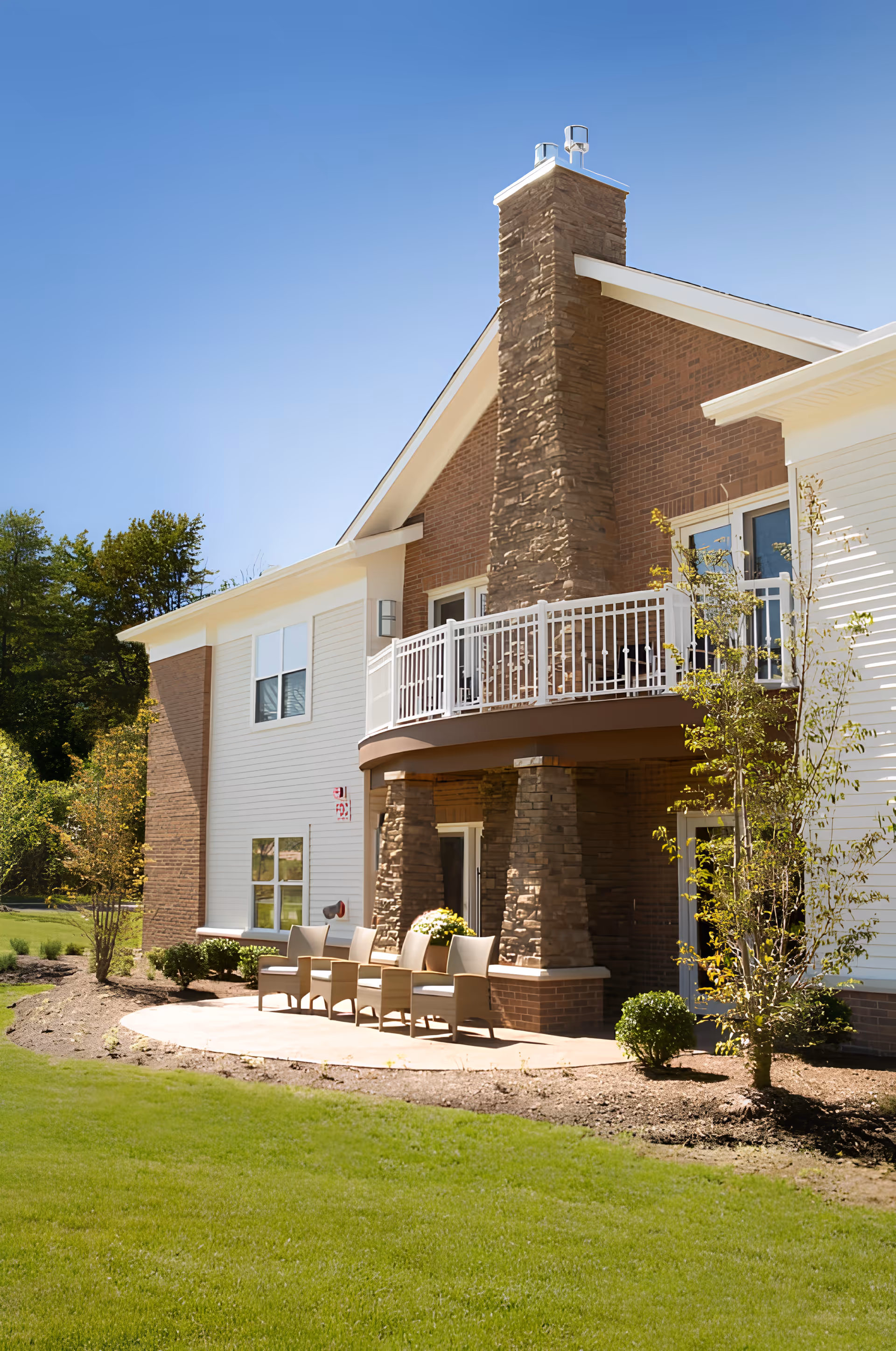 Outdoor patio area of Governor's Village senior living facility featuring a stone chimney, white railing balcony, several chairs arranged on a concrete patio, surrounded by green grass and small trees under a clear blue sky.