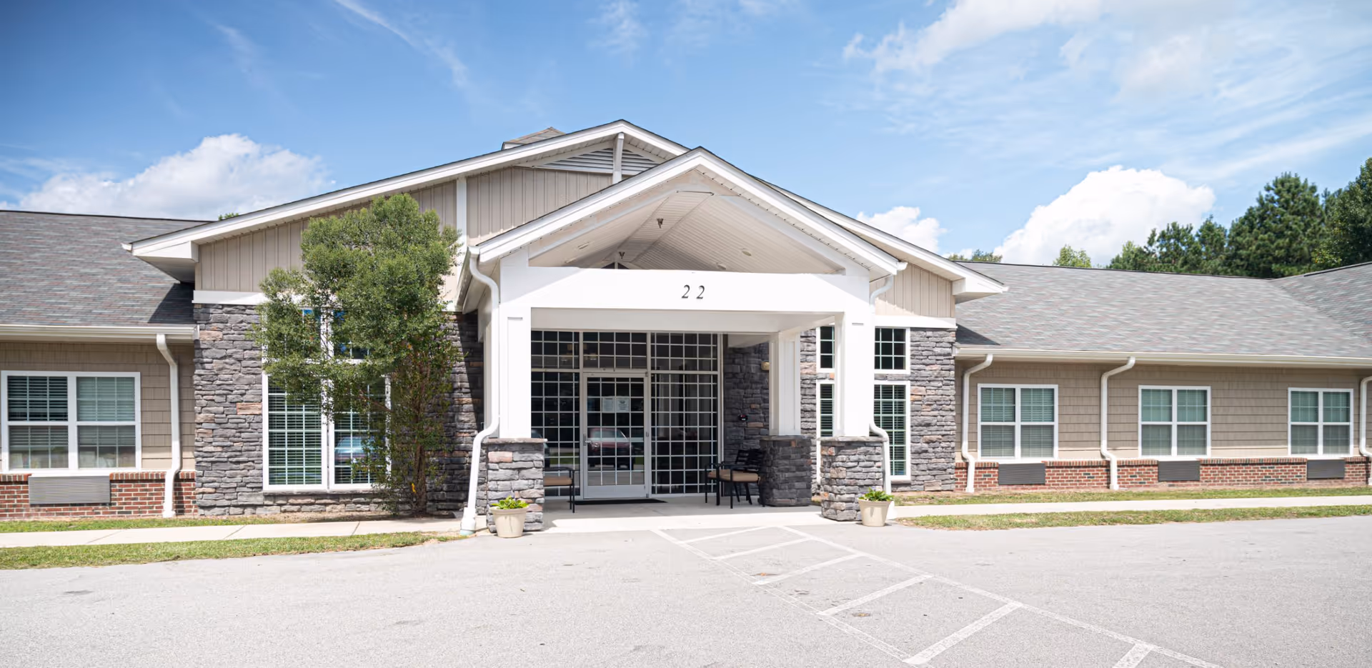 Front exterior view of a single-story building with a covered entrance supported by stone pillars, large windows with white frames, and a sign displaying the number 22 above the entrance. There are two potted plants near the entrance and a clear blue sky with some clouds in the background.