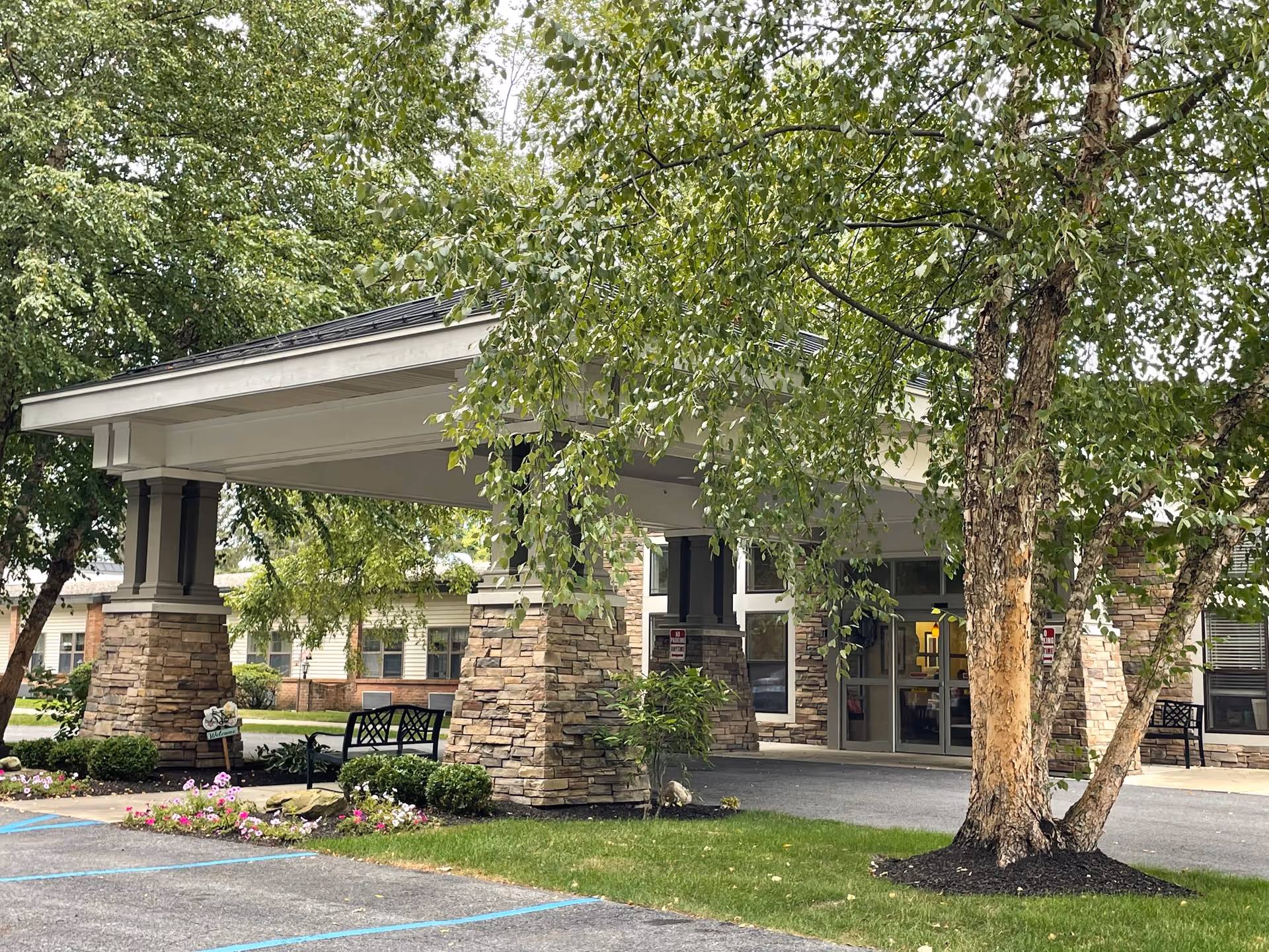 Entrance of Peregrine Senior Living at Guilderland showing a covered drop-off area supported by stone pillars, surrounded by green trees and landscaped flower beds, with benches nearby and a paved driveway.