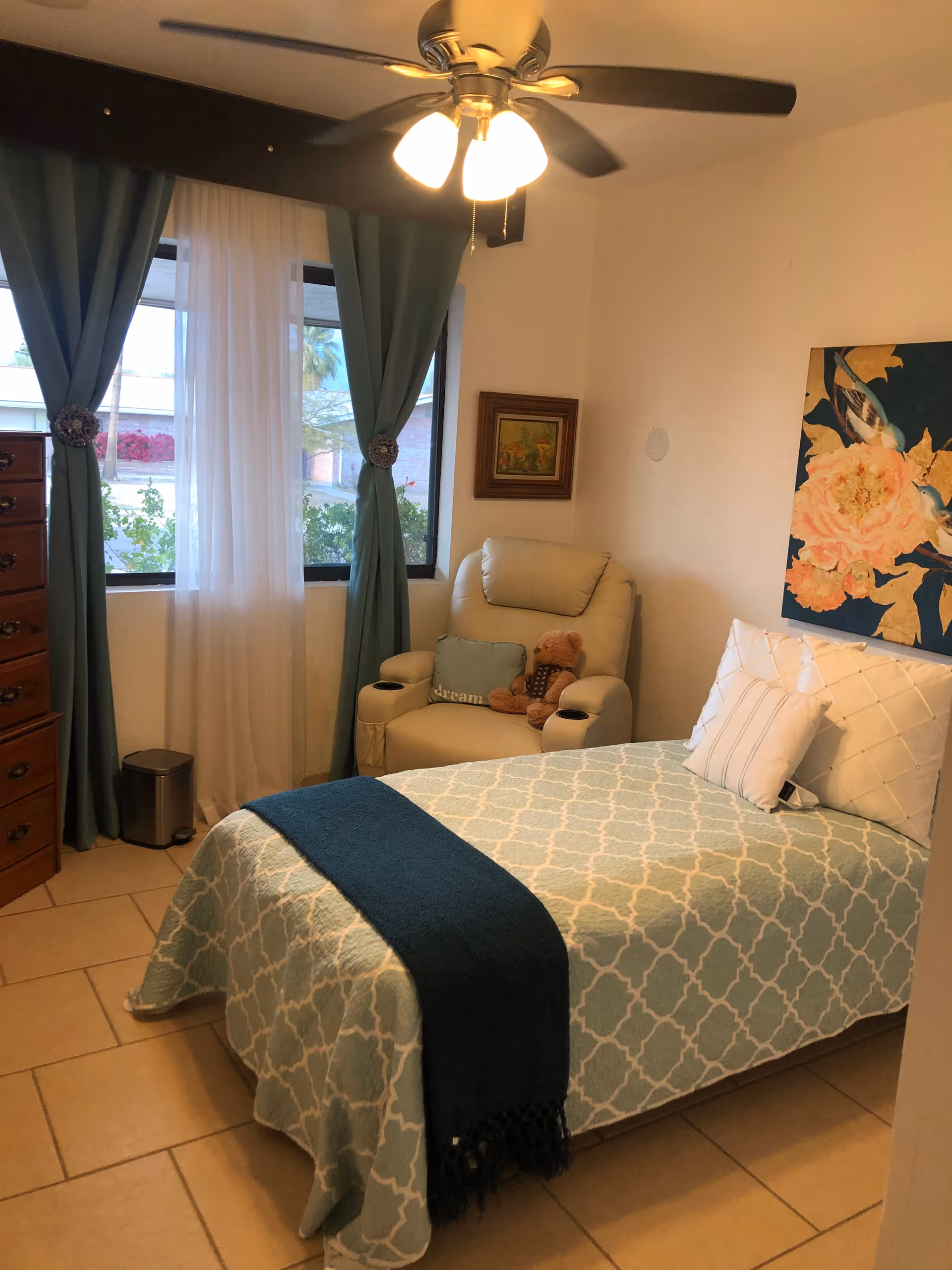 Cozy bedroom in a senior living facility featuring a bed, recliner, dresser, and teal curtains.