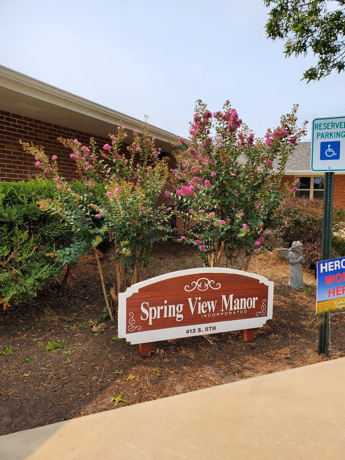 Outdoor view of the entrance area of Spring View Manor Healthcare and Rehabilitation Center featuring a wooden sign with the facility's name and address, surrounded by flowering bushes and greenery. A reserved parking sign for handicapped parking and a small angel statue are also visible nearby.
