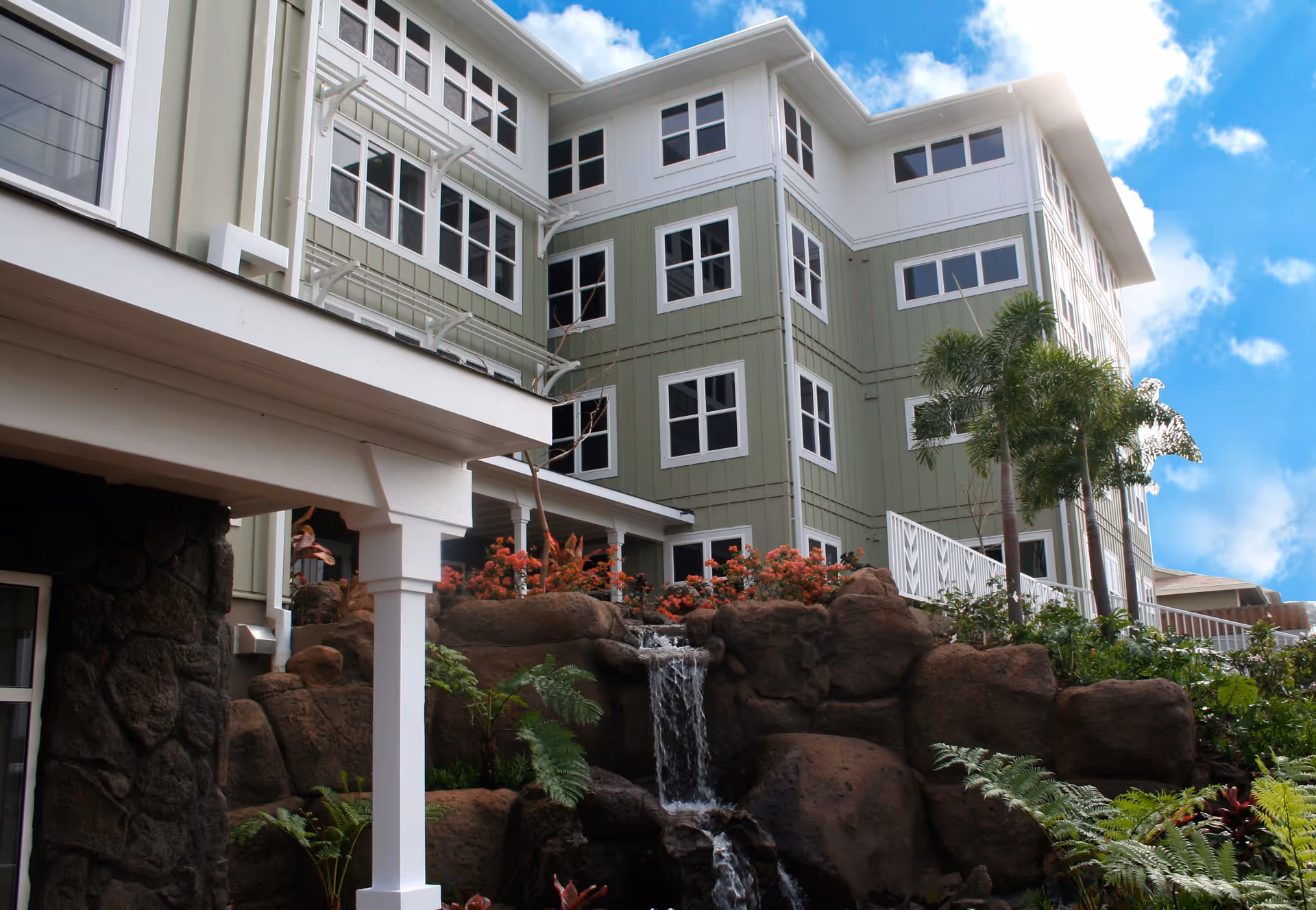 Exterior view of a multi-story senior living facility building with green and white siding, multiple windows, and a landscaped garden featuring a small waterfall and tropical plants under a blue sky with clouds.