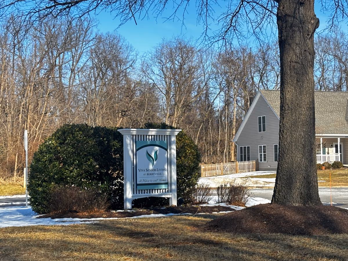 Sign for a senior living facility on a landscaped lawn with a nearby house-like building, bare trees, and a large tree trunk in the foreground.