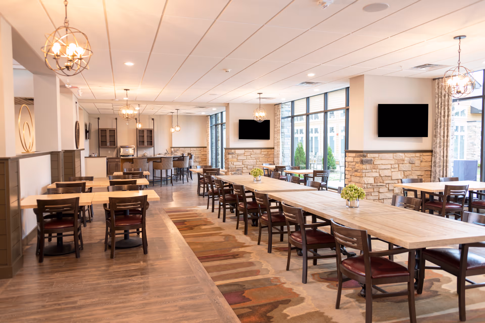 A spacious dining room with multiple wooden tables and chairs arranged neatly. The room features large windows letting in natural light, stone accent walls, and modern hanging light fixtures. There are two flat-screen TVs mounted on the walls and small green plants placed on some tables. The floor has a combination of wood and a patterned rug.
