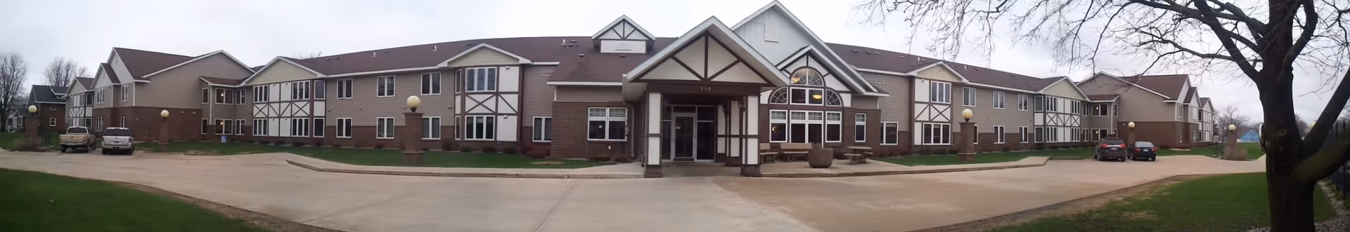 Panoramic exterior front view of a two-story senior living facility with a central entrance, courtyard and parking area.