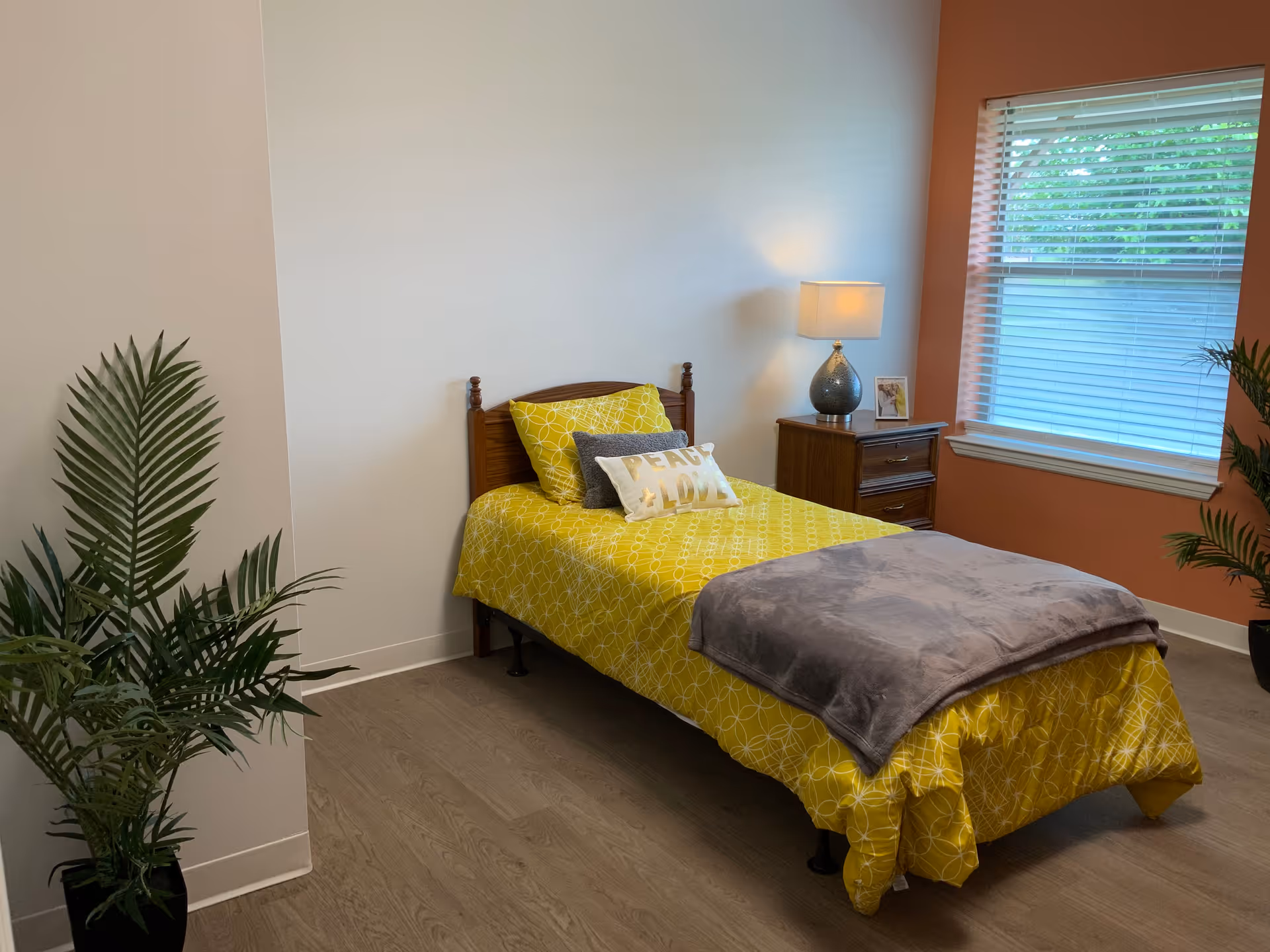 A tidy bedroom with yellow bedding, a wooden bed, nightstand, lamp, and plants.
