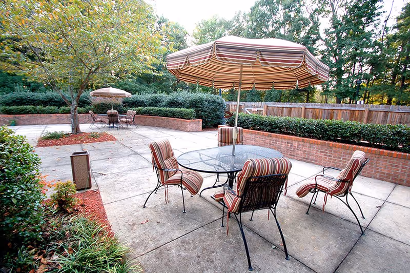 Outdoor patio area with metal tables and chairs featuring striped cushions and matching striped umbrellas. The patio is surrounded by brick planters with green shrubs and trees, and a wooden fence encloses the area.