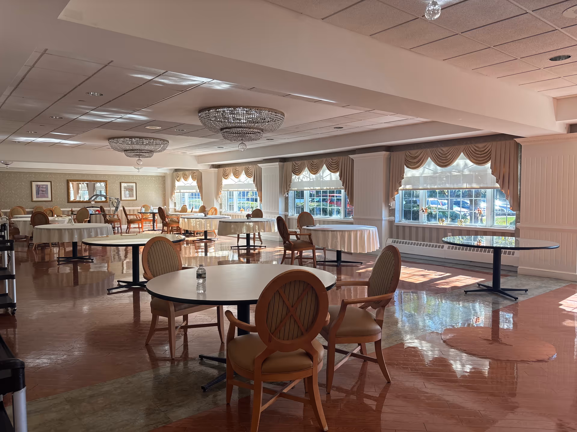 Spacious, empty dining hall with round tables, chairs, chandeliers, and large windows.