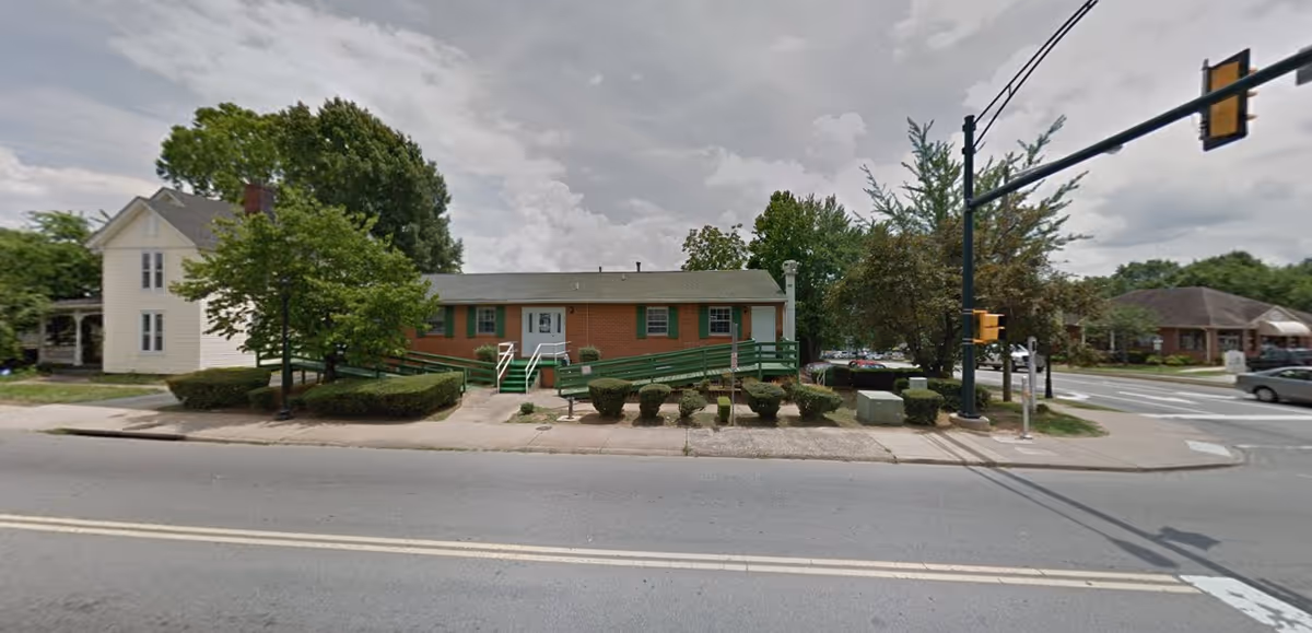 Single-story brick building with green wheelchair ramps and trimmed hedges on a street corner under a cloudy sky.