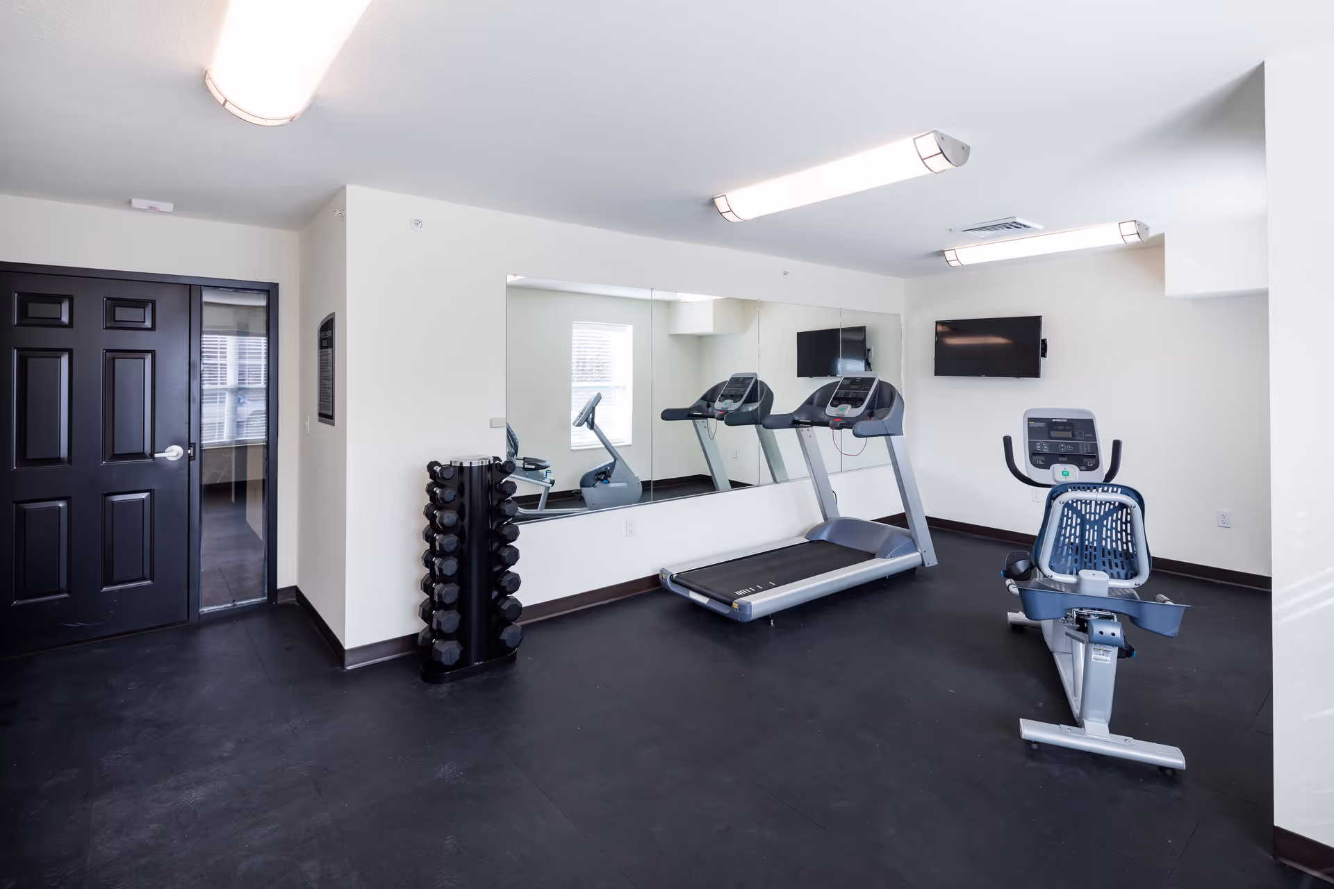 A small fitness room with a treadmill, a recumbent exercise bike, a rack of dumbbells, a wall-mounted TV, and a large mirror on the wall reflecting the equipment and window.