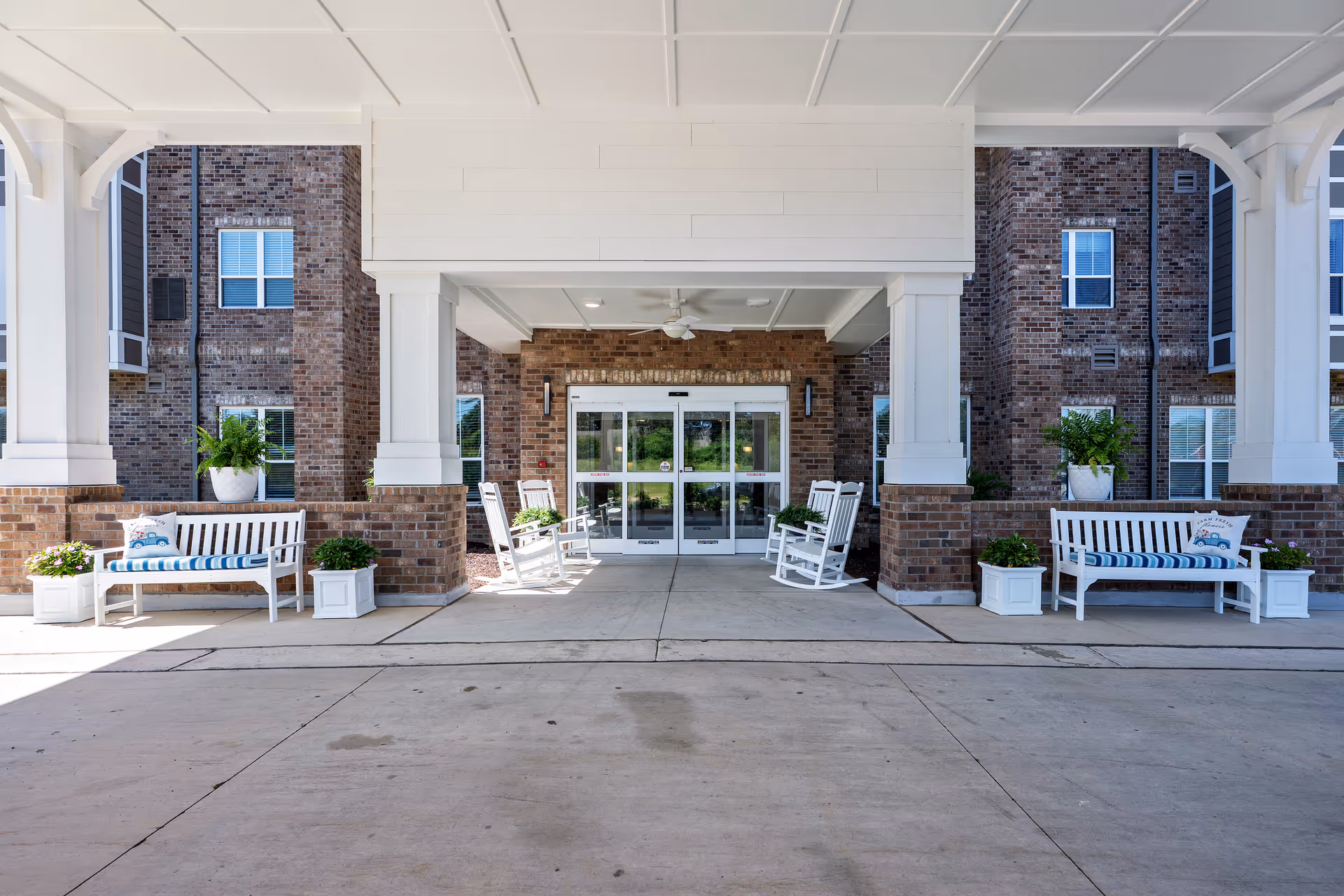 Front entrance of Harmony at Morning Grove with white benches, brick columns, and potted plants.