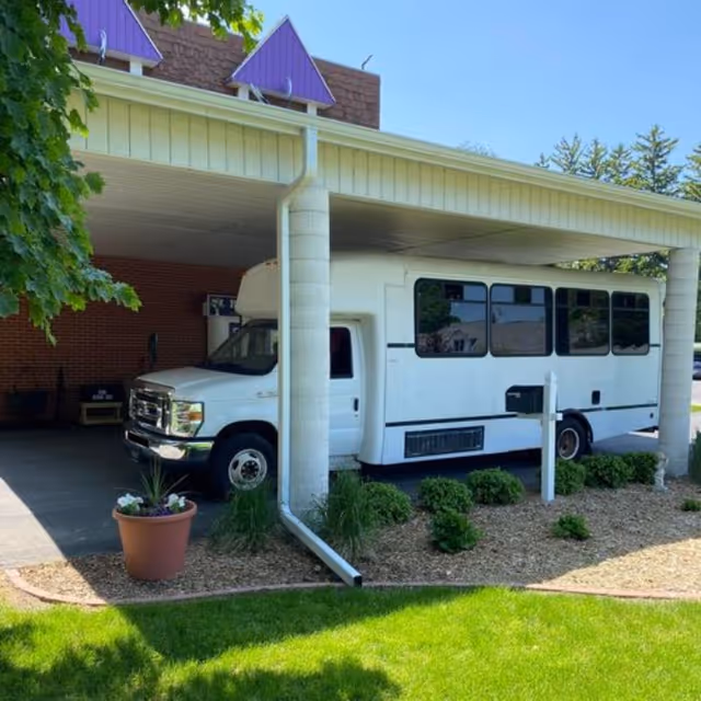 White shuttle bus parked under a covered driveway beside landscaped entrance with potted plants.