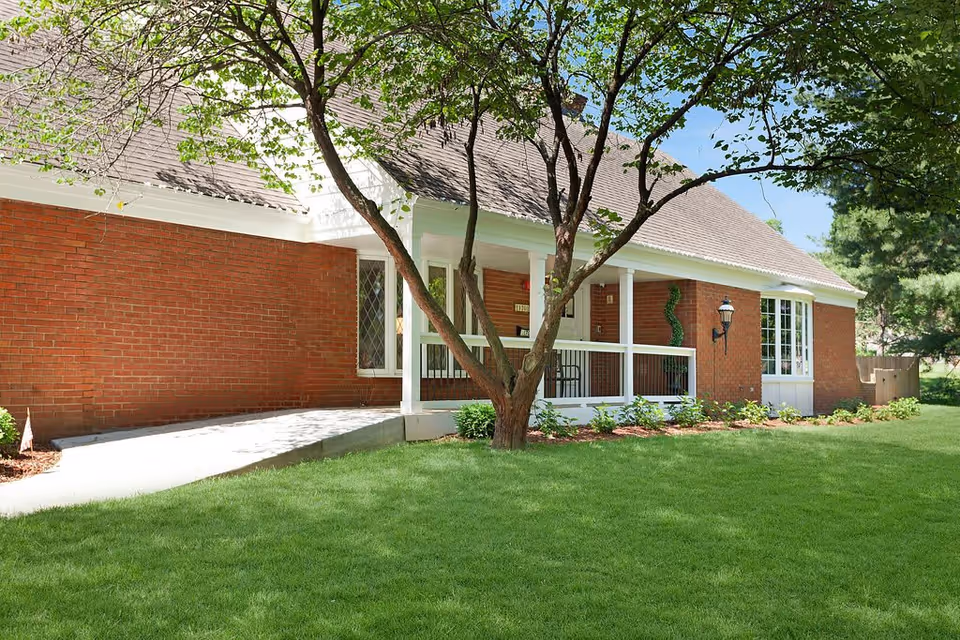 Front of a single-story brick house with a covered porch, tree-shaded lawn, and a concrete walkway.