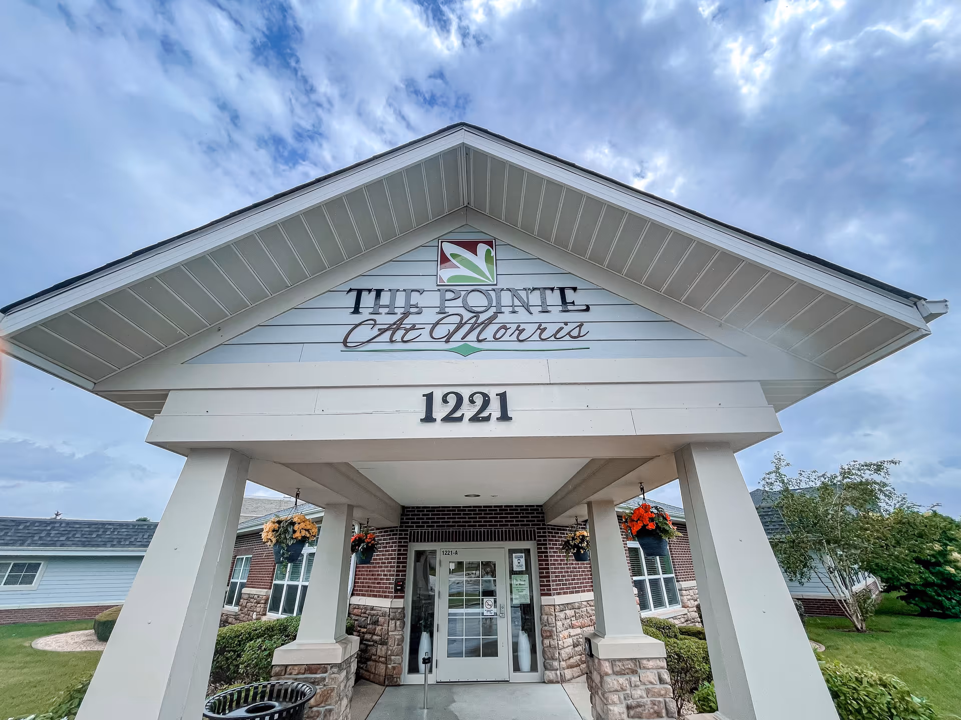 Entrance of The Pointe At Morris facility showing a covered porch with white pillars, hanging flower baskets, and the address number 1221. The building has a brick and stone facade with windows and a glass door under a cloudy sky.