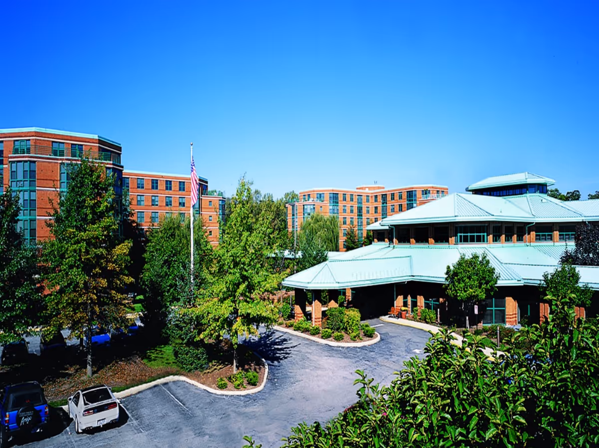 Front exterior view of a multi-wing brick senior living facility with green metal roofs, landscaped grounds, a flagpole, and a parking lot.